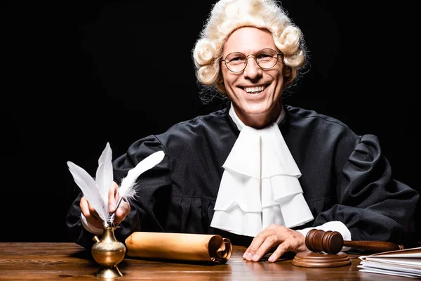Smiling judge in judicial robe and wig taking feather isolated on black — Stock Photo