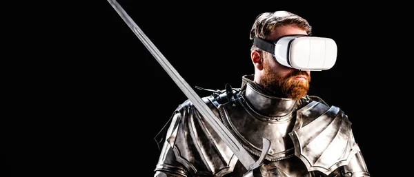 Панорамний знімок лицаря з гарнітурою віртуальної реальності в обладунках, що тримає меч ізольовано на чорному — стокове фото