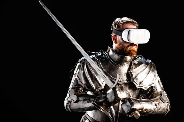 Knight with virtual reality headset in armor holding sword isolated on black — Stock Photo