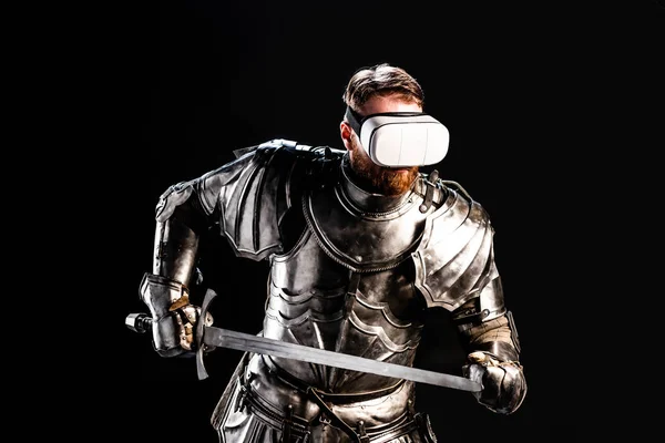 Knight with virtual reality headset in armor holding sword isolated on black — Stock Photo