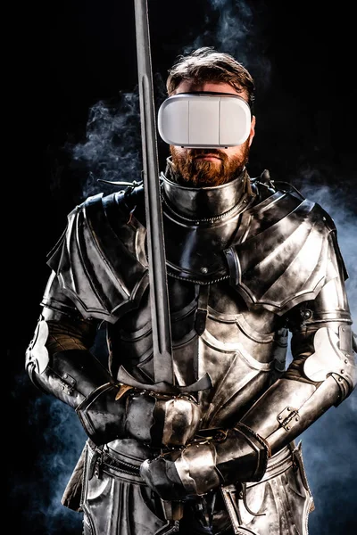Knight with virtual reality headset in armor holding sword on black background — Stock Photo