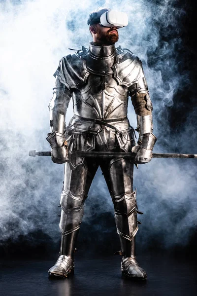 Knight with virtual reality headset in armor holding sword on black background — Stock Photo