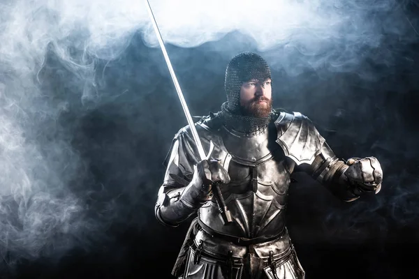 Handsome knight in armor looking away and holding sword on black background — Stock Photo