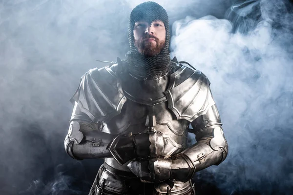 Bonito cavaleiro em armadura olhando para câmera e segurando espada no fundo preto — Fotografia de Stock