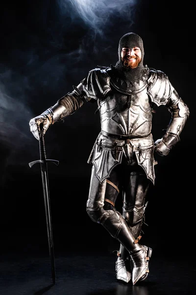 Lächelnder Ritter in Rüstung blickt in die Kamera und hält Schwert auf schwarzem Hintergrund — Stockfoto