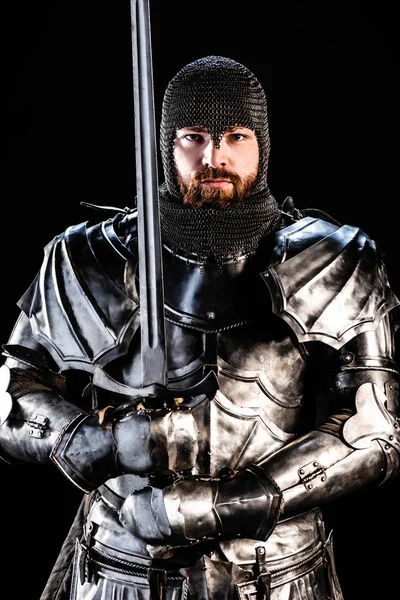 Apuesto caballero de armadura mirando a la cámara y sosteniendo la espada aislada en negro - foto de stock