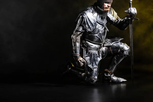 Guapo caballero de armadura sosteniendo la espada y doblar la rodilla sobre fondo negro - foto de stock