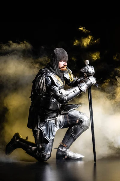 Handsome knight in armor holding sword and bend knee on black background — Stock Photo