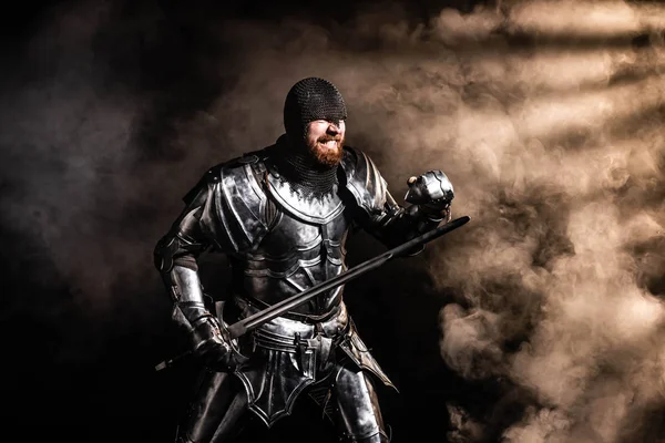 Handsome knight in armor holding sword and fighting on black background — Stock Photo