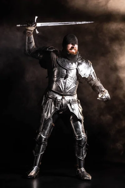 Handsome knight in armor holding sword and fighting on black background — Stock Photo