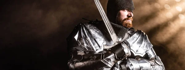 Tiro panorâmico de belo cavaleiro em armadura segurando espada no fundo preto — Fotografia de Stock