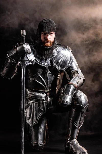 Handsome knight in armor holding sword and bend knee on black background — Stock Photo