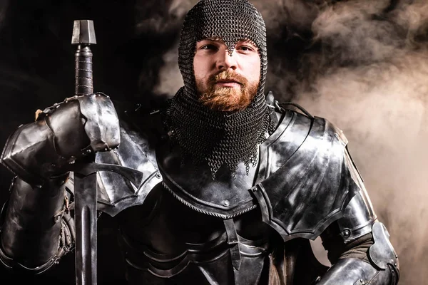 Handsome knight in armor holding sword on black background — Stock Photo