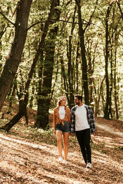 Sonriente joven pareja cogida de la mano y mirándose mientras caminan en el parque - foto de stock