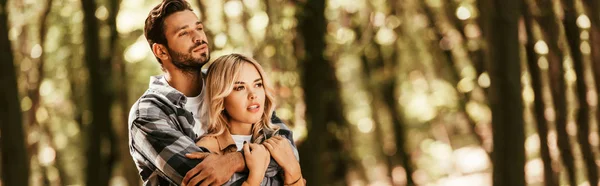 Tiro panorámico de hombre feliz abrazando novia atractiva mientras mira hacia otro lado en el parque - foto de stock