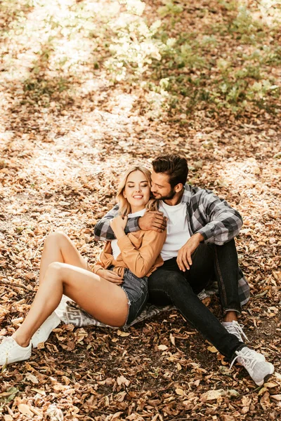 Happy man embracing cheerful girlfriend while sitting on fall foliage in park — Stock Photo