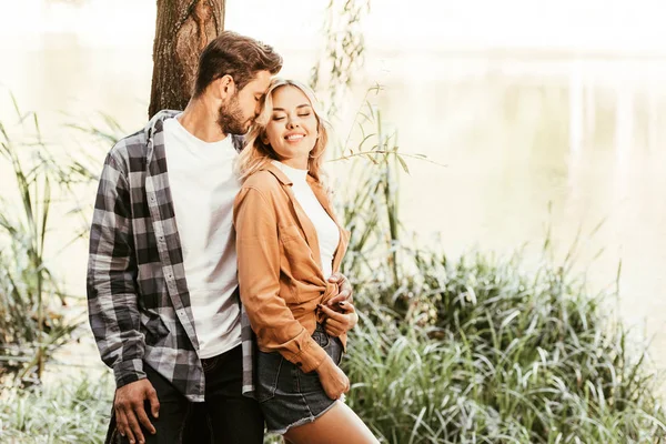 Young man embracing cheerful girlfriend while standing near lake in park — Stock Photo