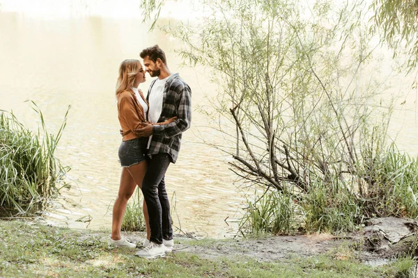 Felice giovane coppia che abbraccia mentre in piedi vicino al lago nel parco — Foto stock