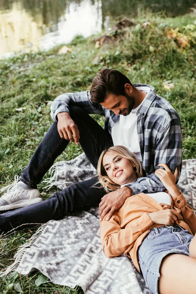 Guapo hombre abrazando feliz novia acostado en manta cerca de lago en parque - foto de stock