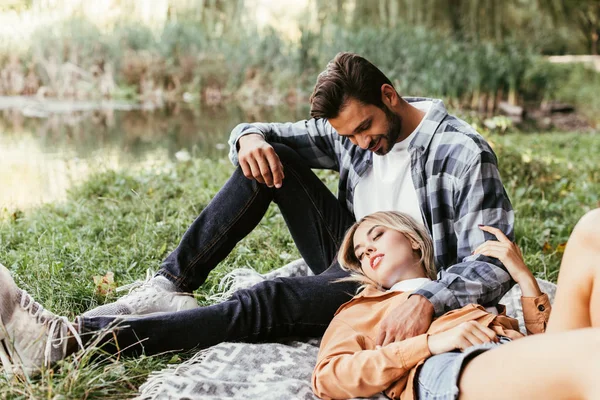 Bello uomo abbracciare fidanzata dormire su coperta vicino lago in parco — Foto stock