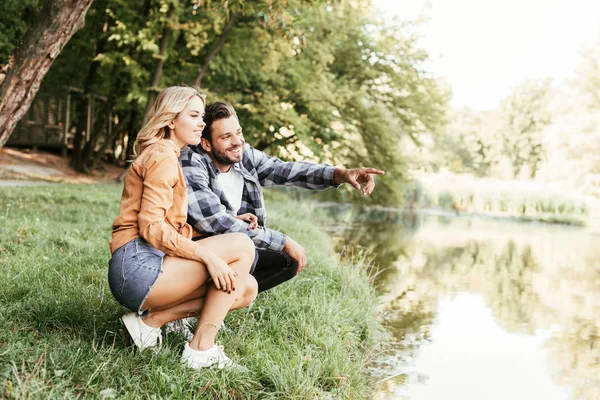 Cheerful man pointing with finger while squatting near lake with girlfriend — Stock Photo