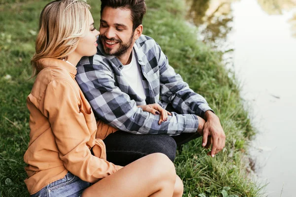 Счастливая молодая пара смотрит друг на друга, сидя на корточках возле озера в парке — стоковое фото