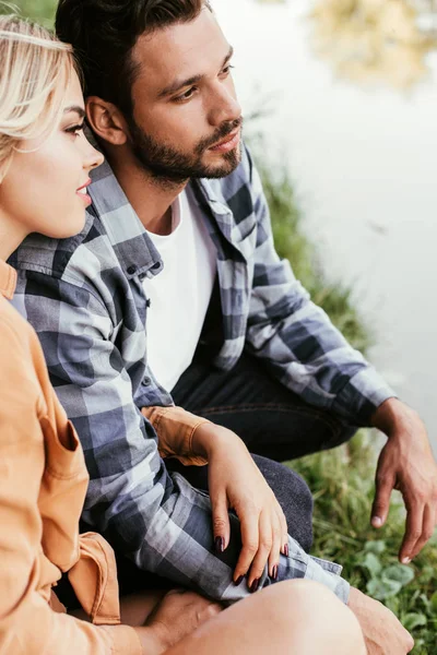 Pareja joven soñadora mirando hacia otro lado mientras pasa tiempo cerca del lago - foto de stock