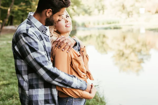 Handsome man embracing happy girlfriend while standing near lake in park — Stock Photo
