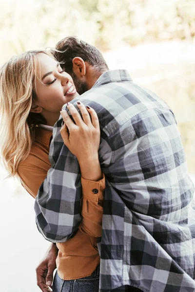 Young man hugging smiling girlfriend while standing near lake in park — Stock Photo