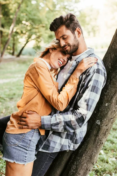 Atractiva mujer joven abrazando novio mientras está de pie cerca del tronco del árbol en el parque - foto de stock