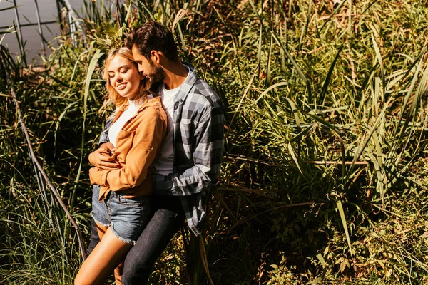 Handsome young man embracing happy girlfriend in thicket of sedge near lake — Stock Photo