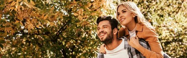 Panoramic shot of cheerful man piggybacking happy girlfriend in park — Stock Photo