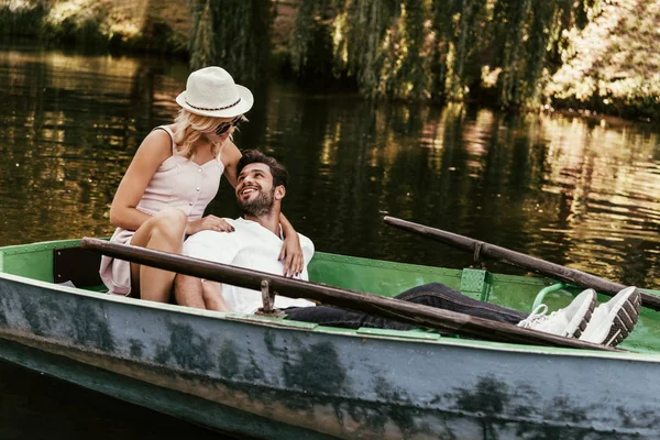 Jovem mulher em sundress e chapéu abraçando namorado feliz enquanto sentado em barco — Fotografia de Stock