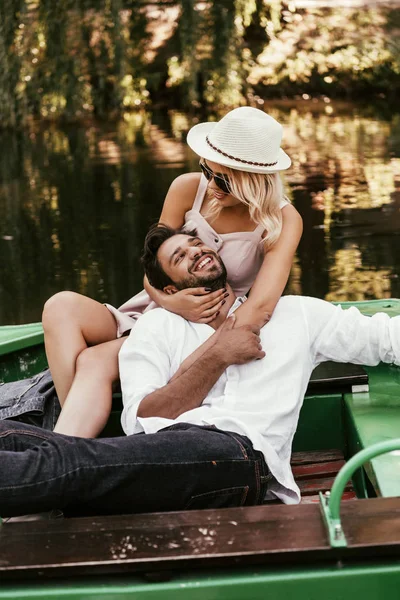Young woman in hat and sunglasses hugging happy boyfriend while sitting in boat — Stock Photo