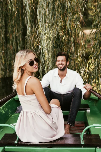 Привлекательная молодая женщина смотрит в камеру, сидя в лодке рядом с счастливым парнем — стоковое фото