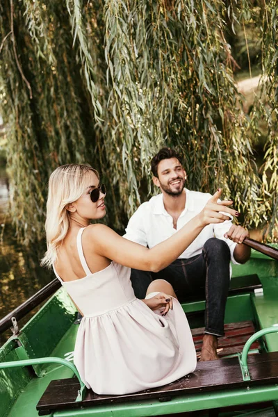 Atractiva mujer joven señalando con el dedo mientras está sentado en el barco cerca de novio feliz - foto de stock