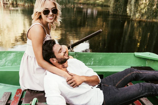 Chica atractiva en gafas de sol abrazando novio feliz mientras está sentado en el barco en el lago - foto de stock
