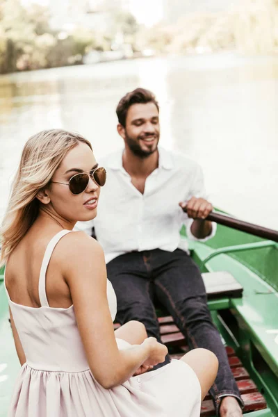 Attractive girl in sunglasses looking at camera while sitting in boat with boyfriend — Stock Photo