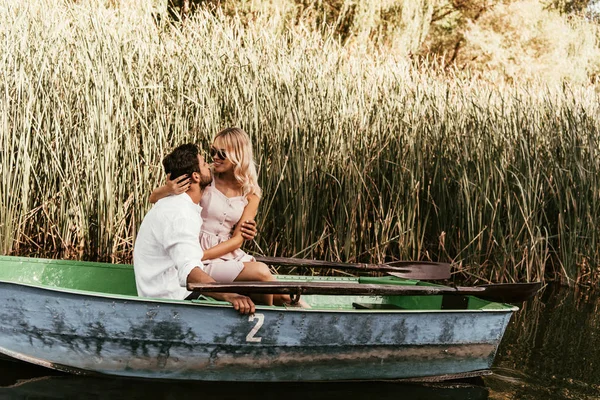 Young couple hugging and kissing in boat on river near thicket of sedge — Stock Photo