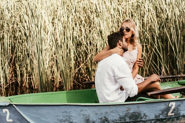 Attractive young woman embracing boyfriend in boat on lake near thicket of sedge — Stock Photo