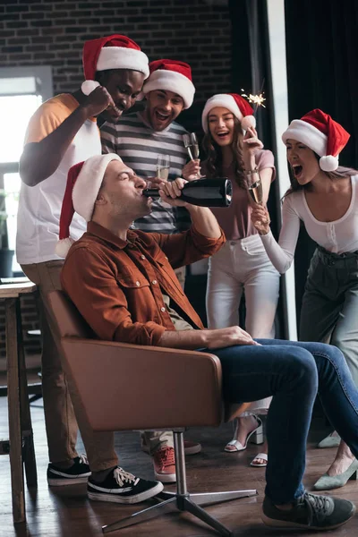 Joven hombre de negocios sentado en sillón y beber champán de la botella cerca de compañeros multiculturales alegres en sombreros de santa - foto de stock