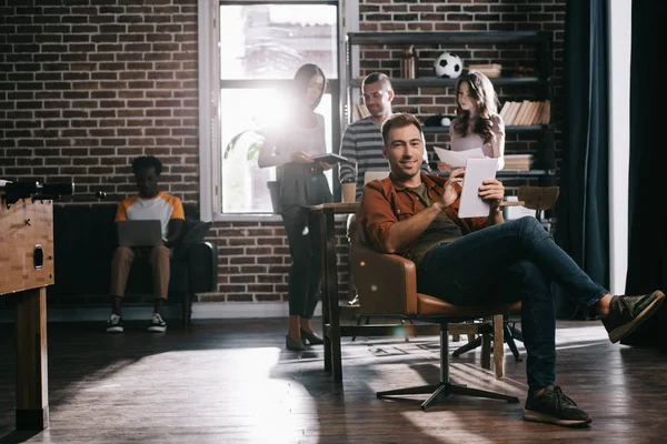 Joven y sonriente hombre de negocios sentado en sillón con tableta digital cerca de colegas multiculturales - foto de stock