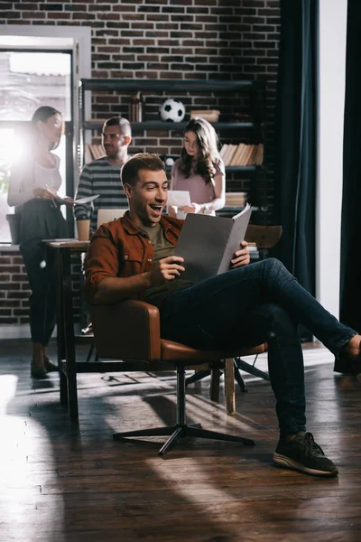 Веселий бізнесмен сидить у кріслі і читає документ біля молодих колег — стокове фото