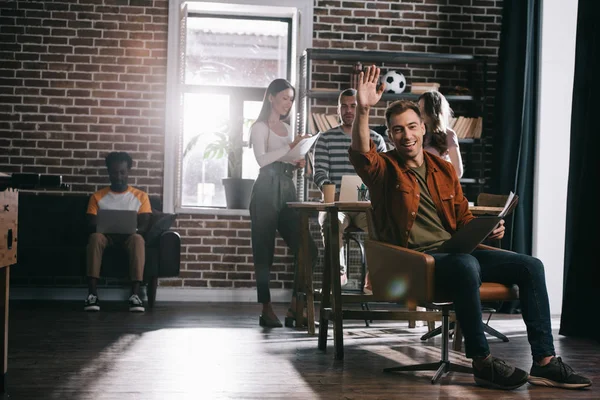 Cheerful businessman sitting in armchair and waving hand at camera near young multicultural colleagues — Stock Photo