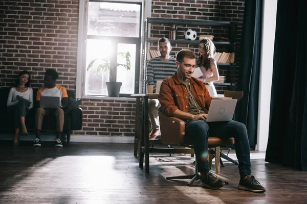 Sonriente hombre de negocios sentado en el sillón y el uso de la computadora portátil cerca de jóvenes colegas multiculturales - foto de stock