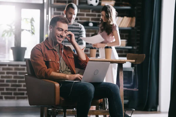 Uomo d'affari sorridente seduto in poltrona, utilizzando laptop e ascoltando musica vicino a giovani colleghi — Foto stock