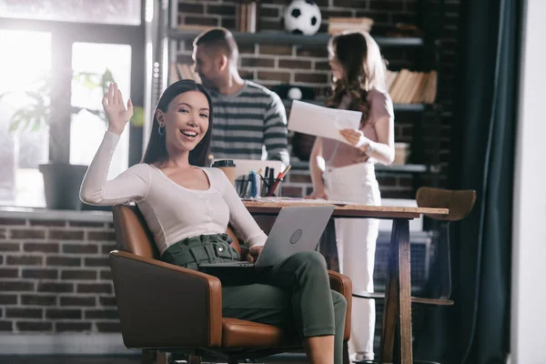 Junge Geschäftsfrau sitzt mit Laptop im Sessel und winkt neben Kollegen in die Kamera — Stockfoto