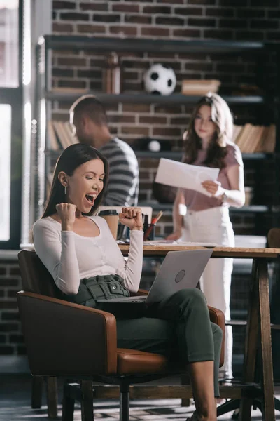 Joven empresaria sentada en sillón con portátil y mostrando el gesto ganador cerca de colegas - foto de stock