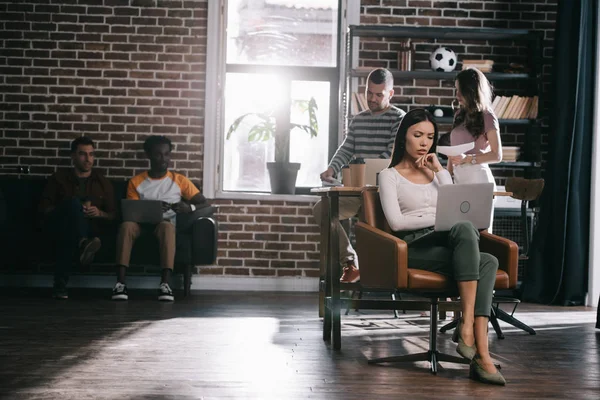 Thoughtful businesswoman sitting in armchair and using laptop near young multicultural colleagues — Stock Photo