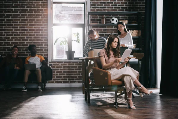 Концентрована бізнес-леді сидить у кріслі і використовує цифровий планшет біля молодих багатокультурних колег — стокове фото